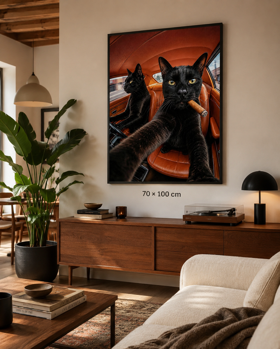 A framed wall art print showing two black cats inside a warm orange-brown car, displayed above a wooden console in a styled living room. The print includes a size marker reading 70 x 100 cm.