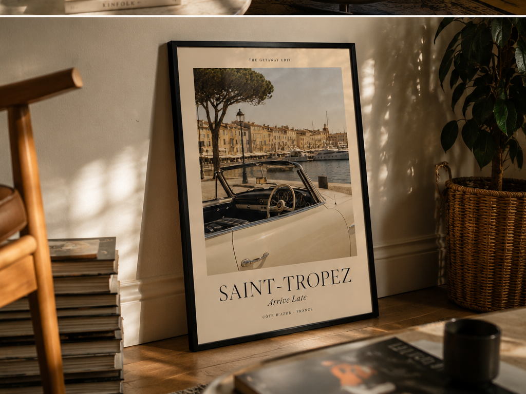 A framed travel poster of Saint-Tropez, France, leaning against a wall on a wooden shelf in a cozy room setting with books and a potted plant.