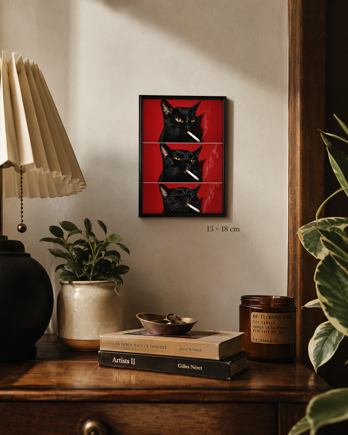A framed wall art print showing two black cats against a red background, displayed in a styled room above a wooden table with books, a plant, and a lamp. The print includes size text reading 13 x 18 cm.