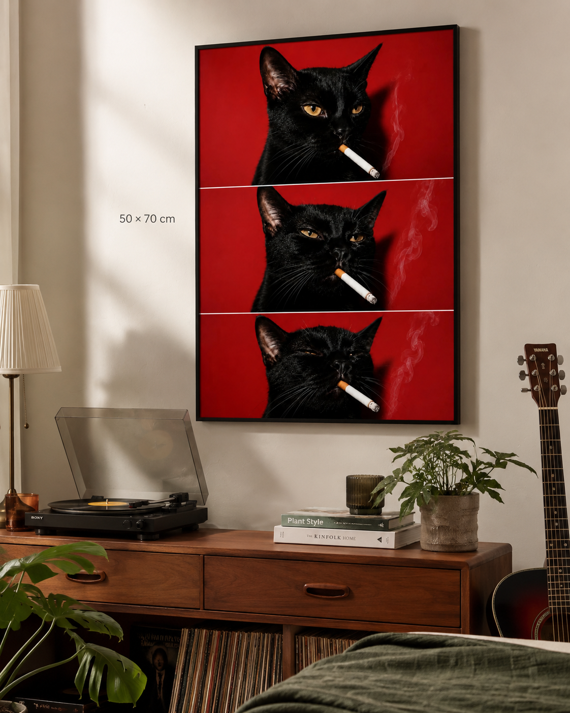 A framed wall art print showing a black cat with a cigarette in three stacked panels on a red background, displayed above a wooden console table in a styled room. The print includes a small '50 x 70 cm' size marker.